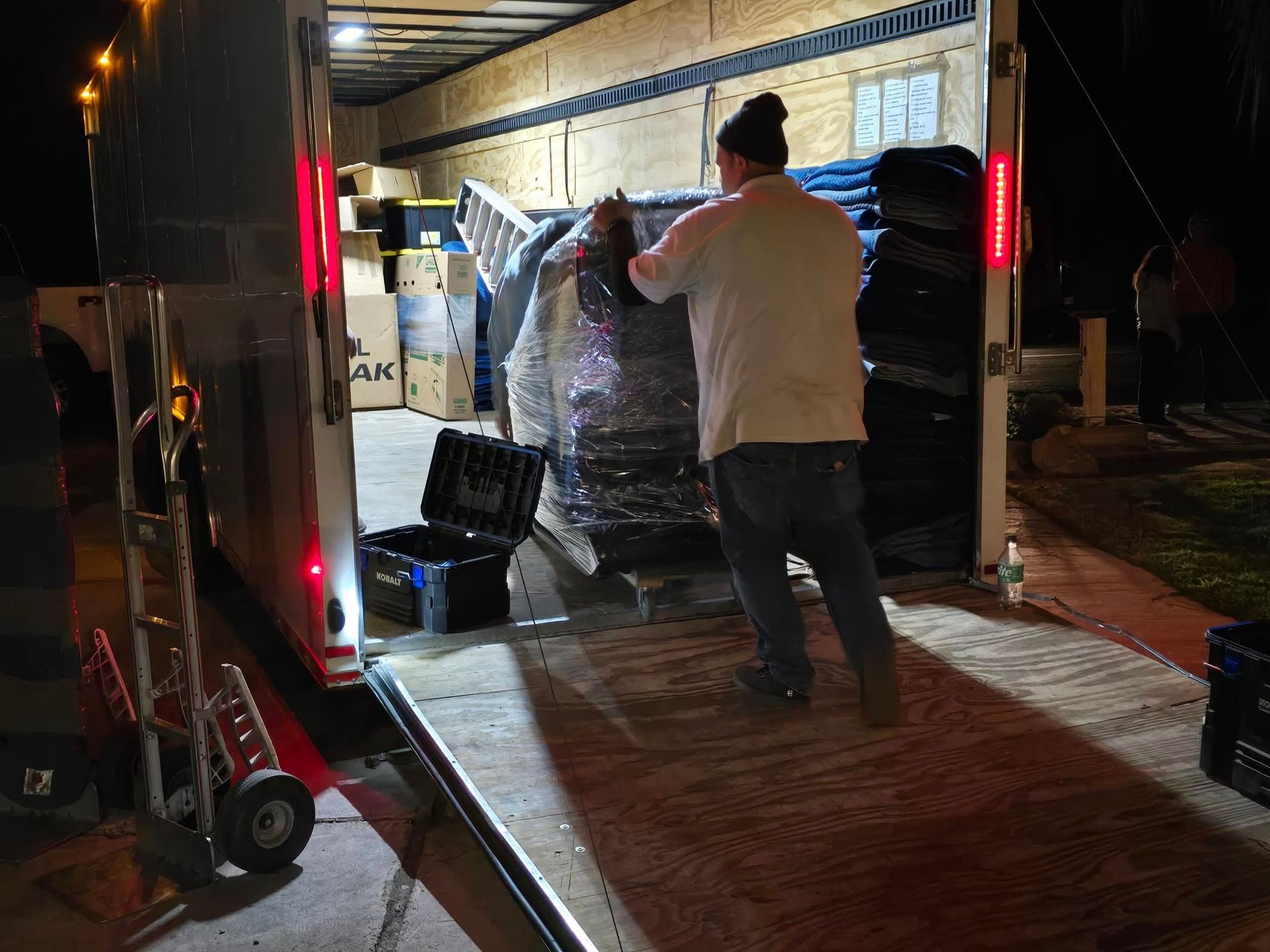 A person in a light shirt and jeans loads a plastic-wrapped item into a brightly lit moving truck at night.