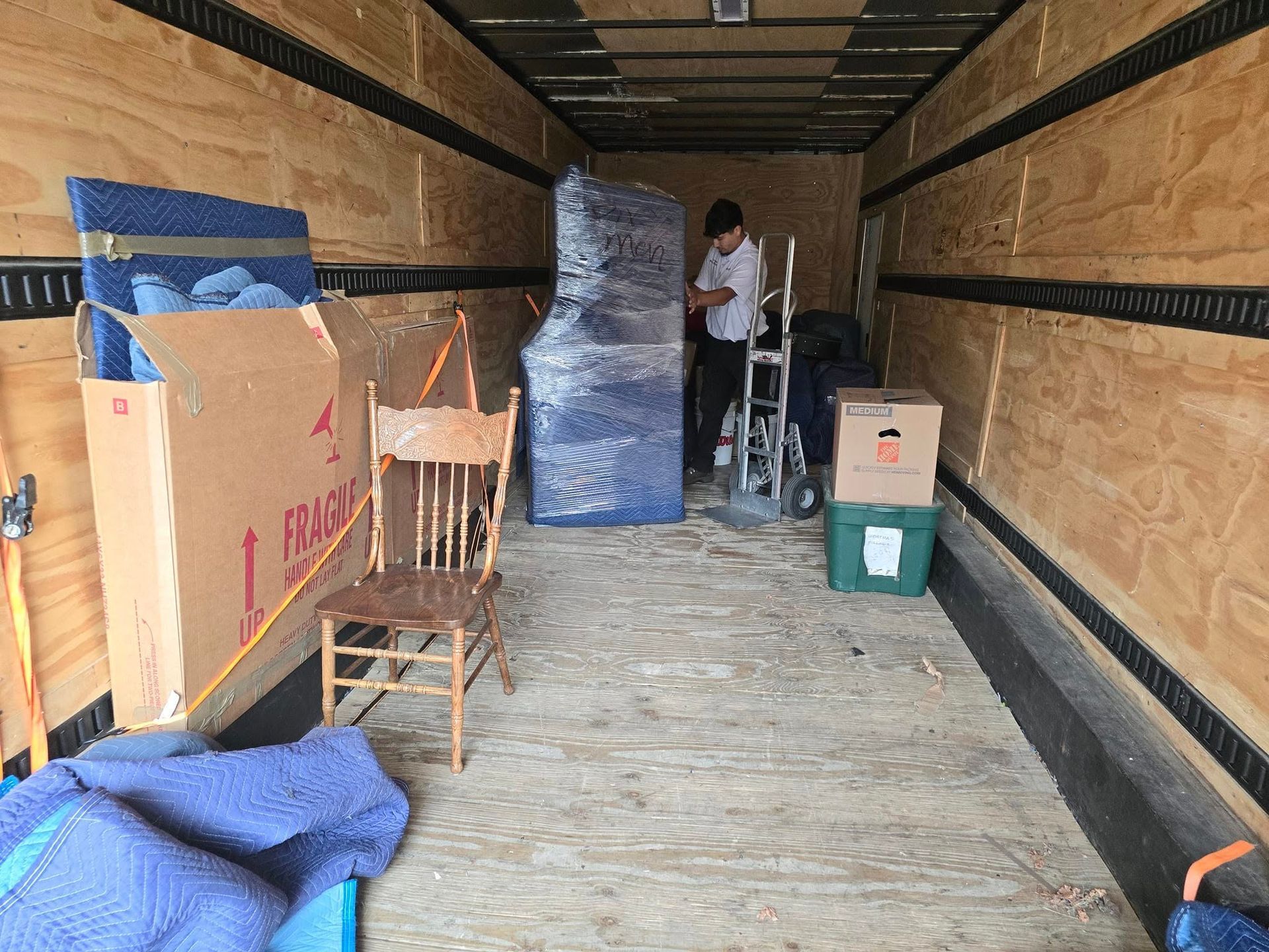 A person stands inside a moving truck, next to a wrapped furniture item, a wooden chair, and several moving boxes.