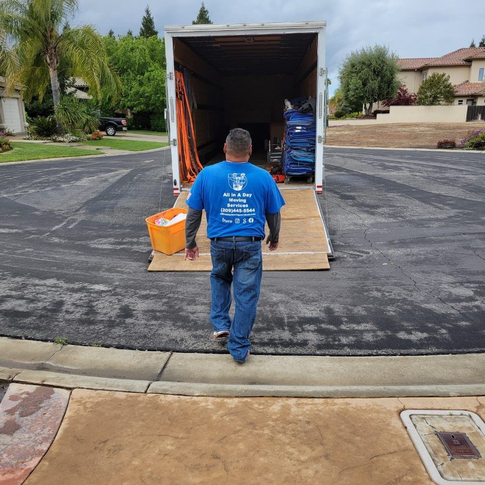 A person wearing a blue shirt walks toward the open back of a moving truck parked on a suburban street.