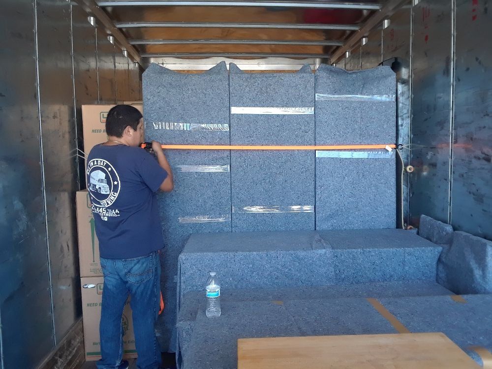 A person in a blue t-shirt tightens a yellow ratchet strap across furniture items wrapped in grey moving blankets.