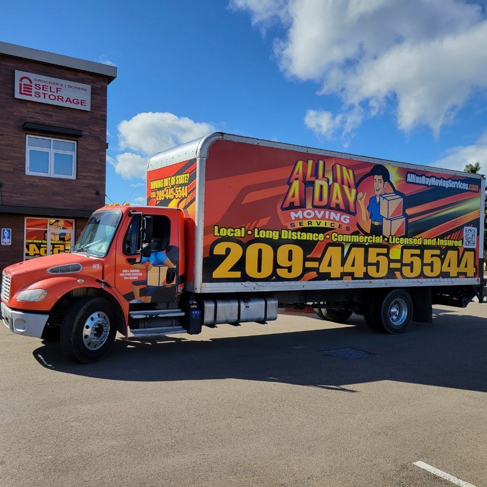 An orange All In A Day Moving truck parked in front of a brick storage facility under a bright blue sky.