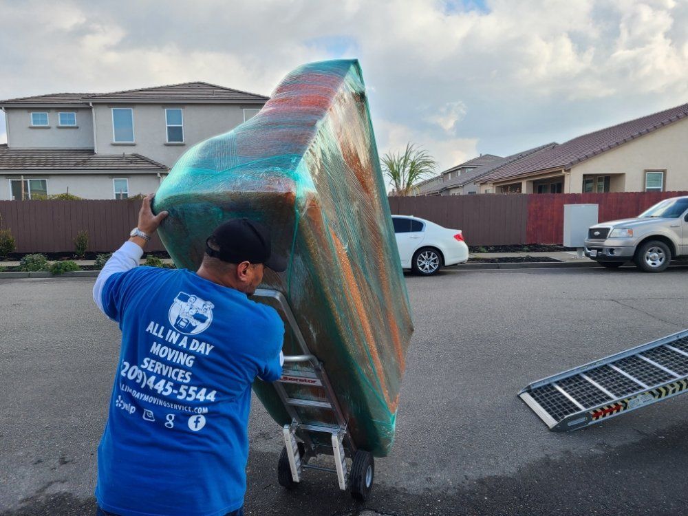 A mover in a blue shirt pushes a piano on a dolly toward a ramp in a residential street.