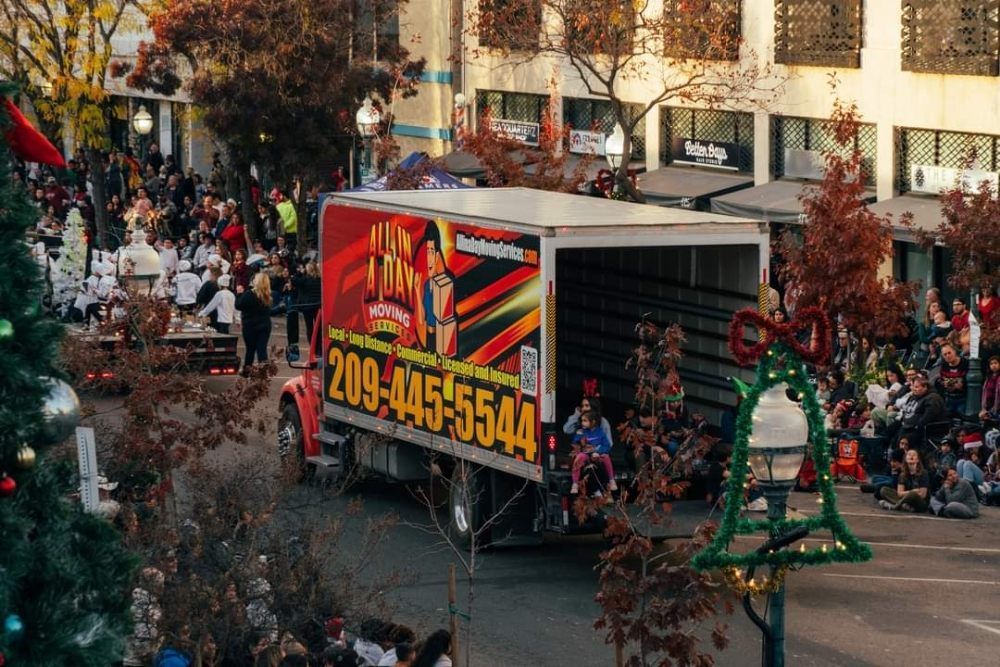 A box truck decorated for a parade drives through a crowd-lined street next to a decorated Christmas tree and bell.