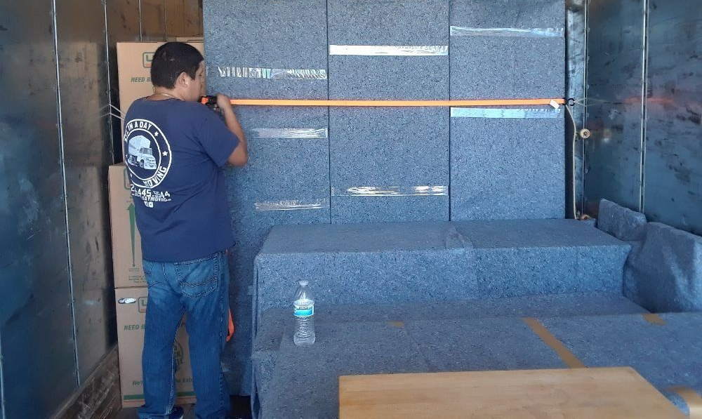 A person in a blue shirt uses a tape measure to measure a wall of large, grey-padded objects inside a moving truck.