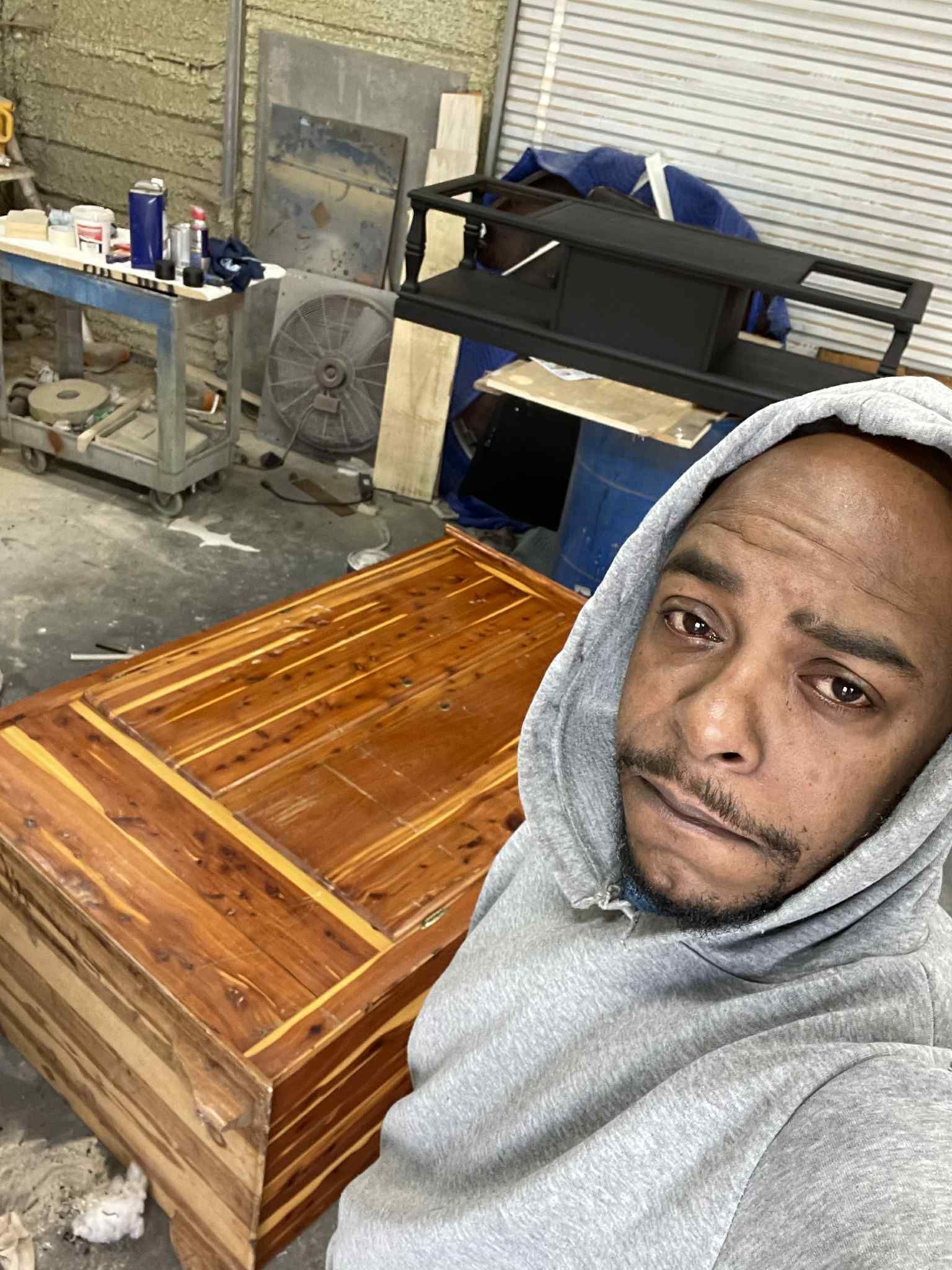 Man in gray hoodie with wooden chest in workshop.
