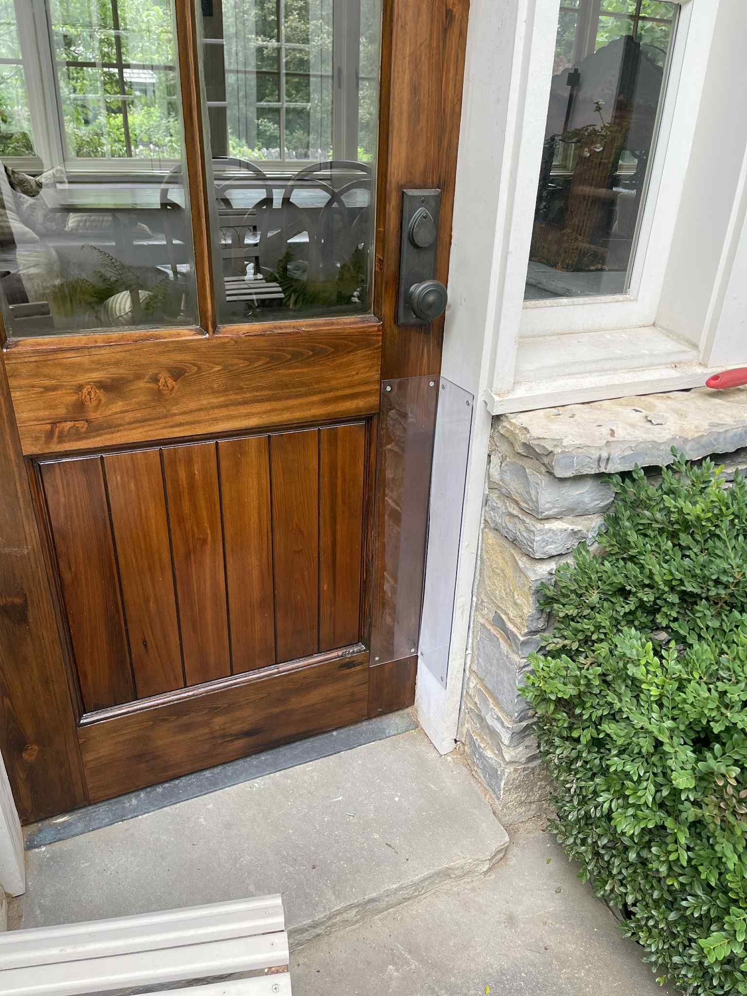 Wooden front door with glass panel, next to stone wall and shrub.
