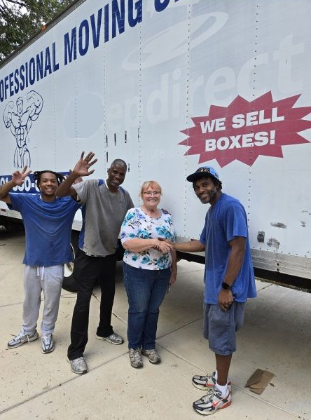 Four people pose by a moving truck. Two men wave, a woman smiles, and another man points.