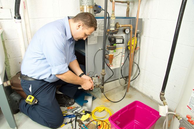 A person kneels, working on a heating system in a basement; tools and pink bin in view.