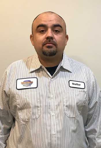 Man wearing a striped work shirt with a name tag. Standing in front of a neutral background.