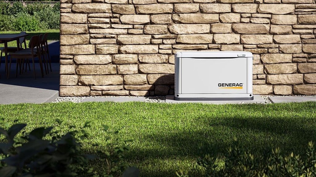 A generator is sitting in the grass next to a brick wall.