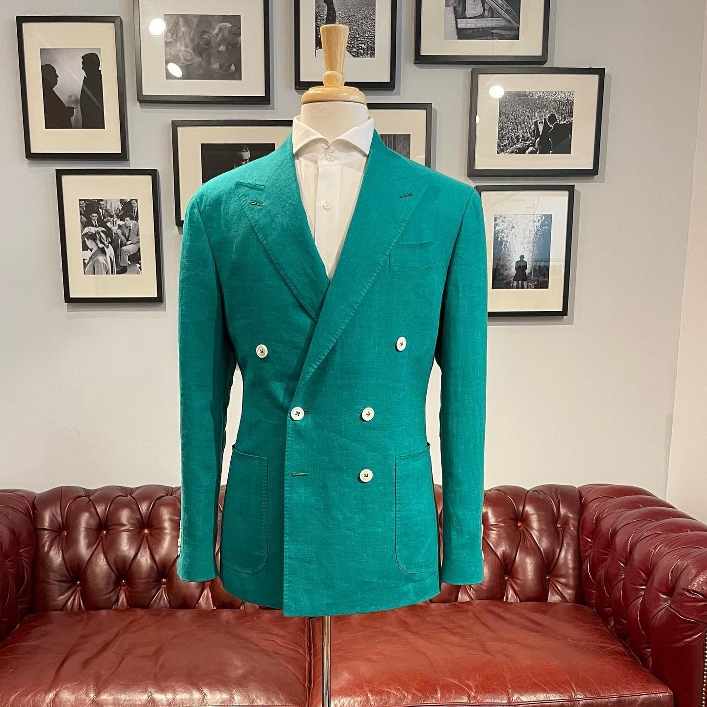 A bright teal, double-breasted textured blazer on a mannequin, set against a wall with framed photos and a leather sofa.