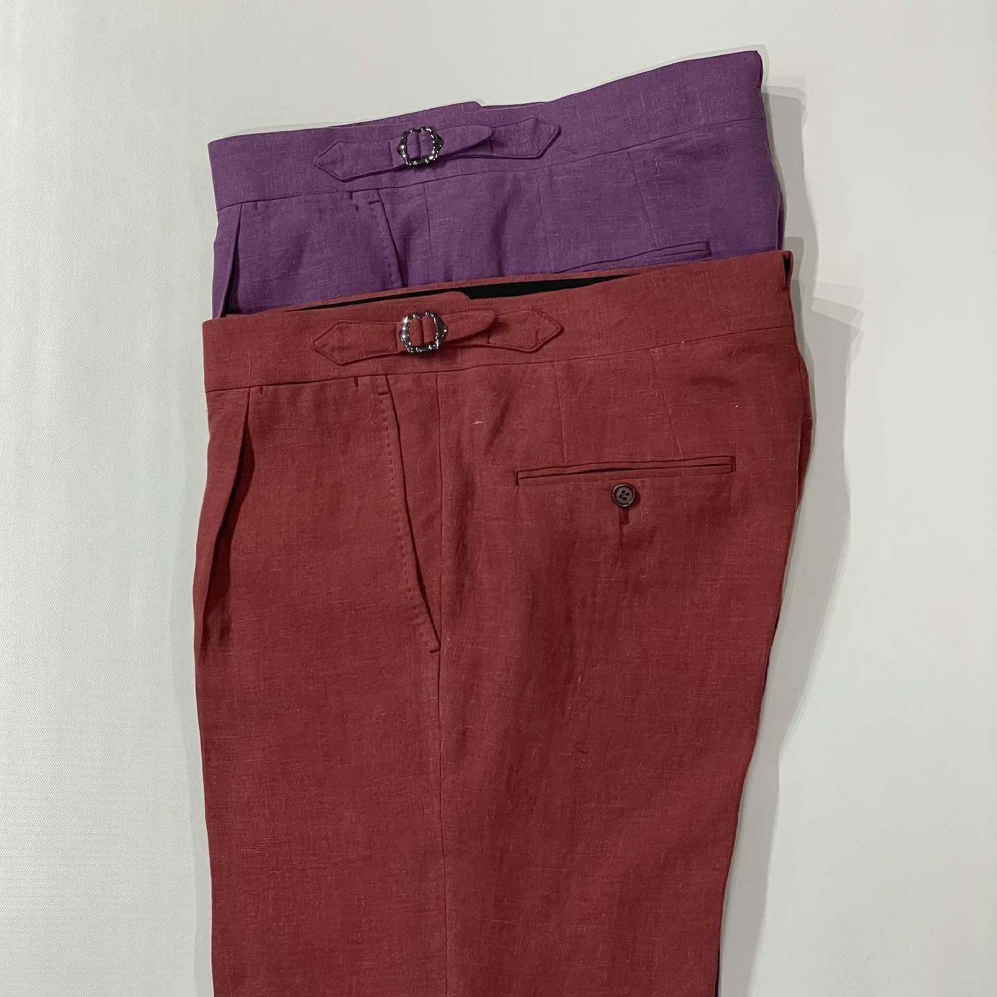 A pair of reddish-brown linen trousers stacked on top of a pair of purple linen trousers with side-adjuster buckles.