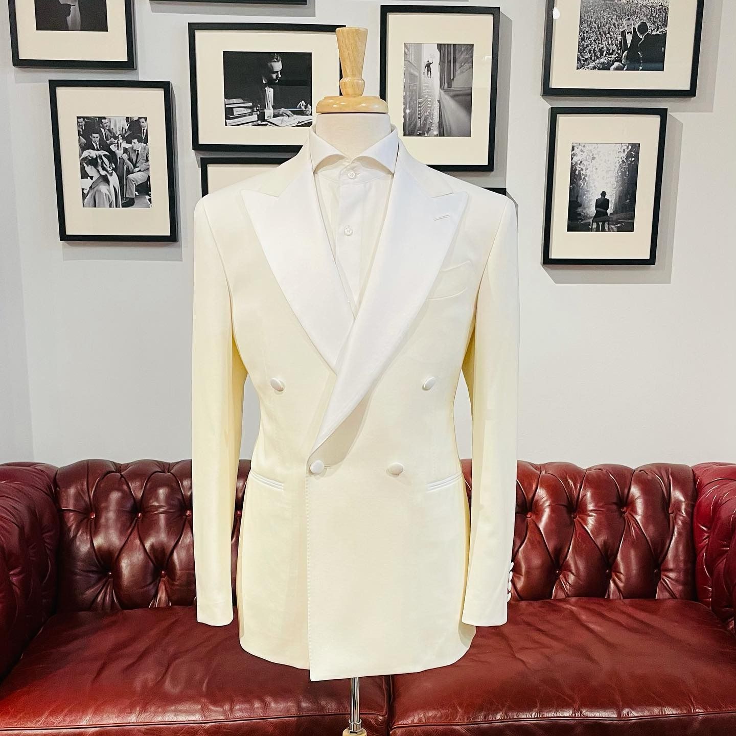 A double-breasted cream tuxedo jacket with white satin lapels on a mannequin, set before a red leather sofa and wall art.