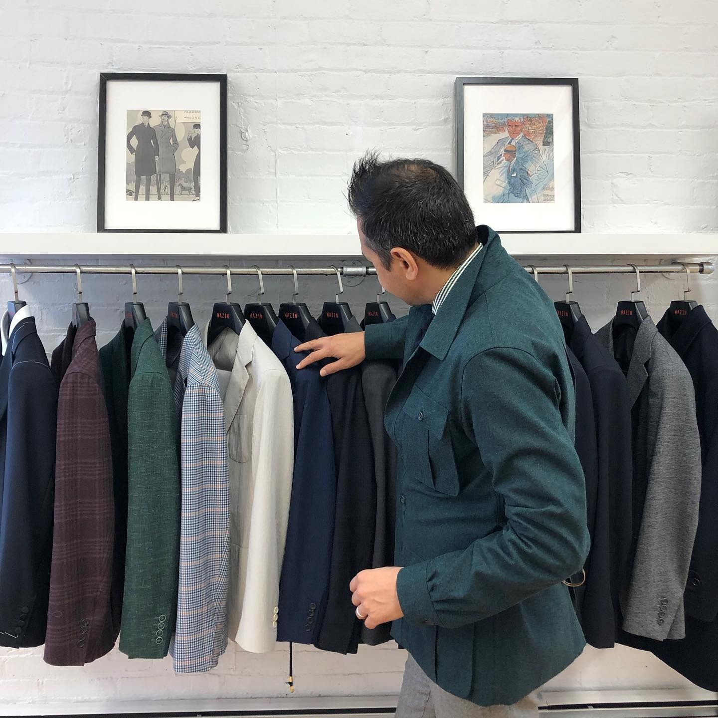 A person in a dark green jacket browsing a rack of assorted blazers and coats displayed in a store with framed art.