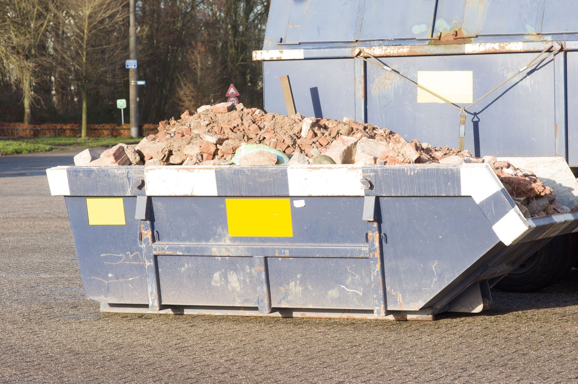 A blue-gray dumpster filled with rubble and debris on a paved lot outdoors.