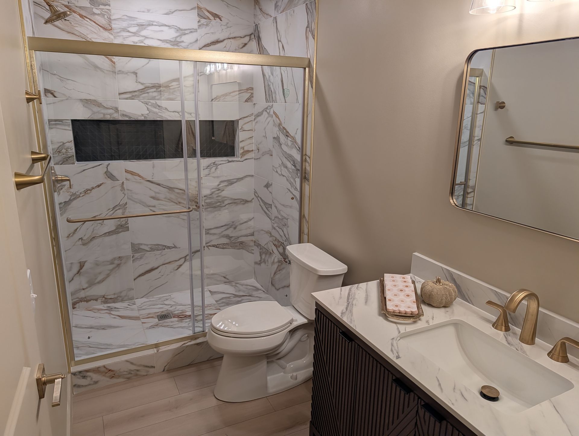 Bathroom with marble-like shower tile, gold fixtures, toilet, vanity with sink, and mirror.