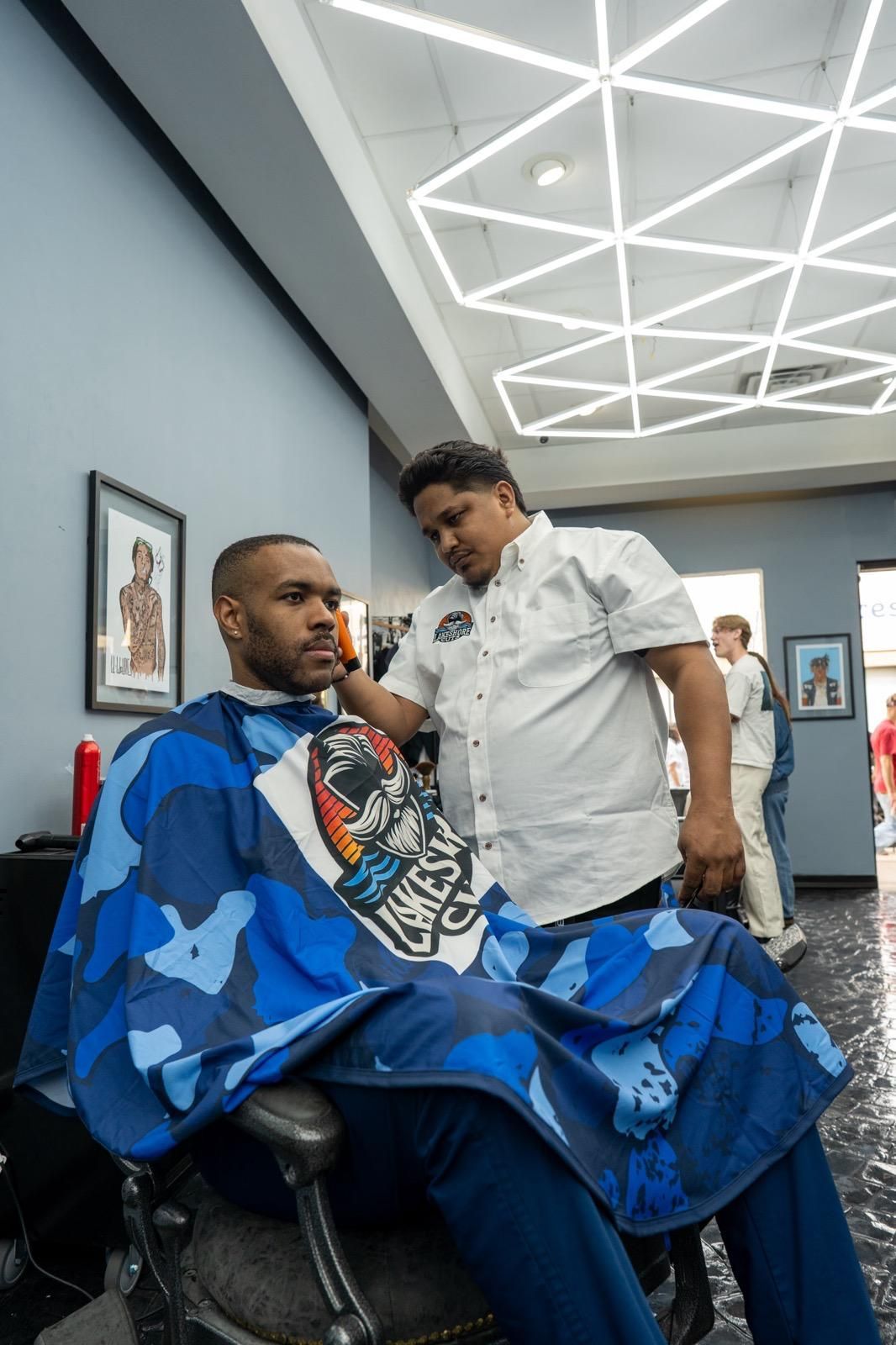 Barbershop scene: a barber trims a seated client draped in a blue cape, with geometric ceiling lights overhead.