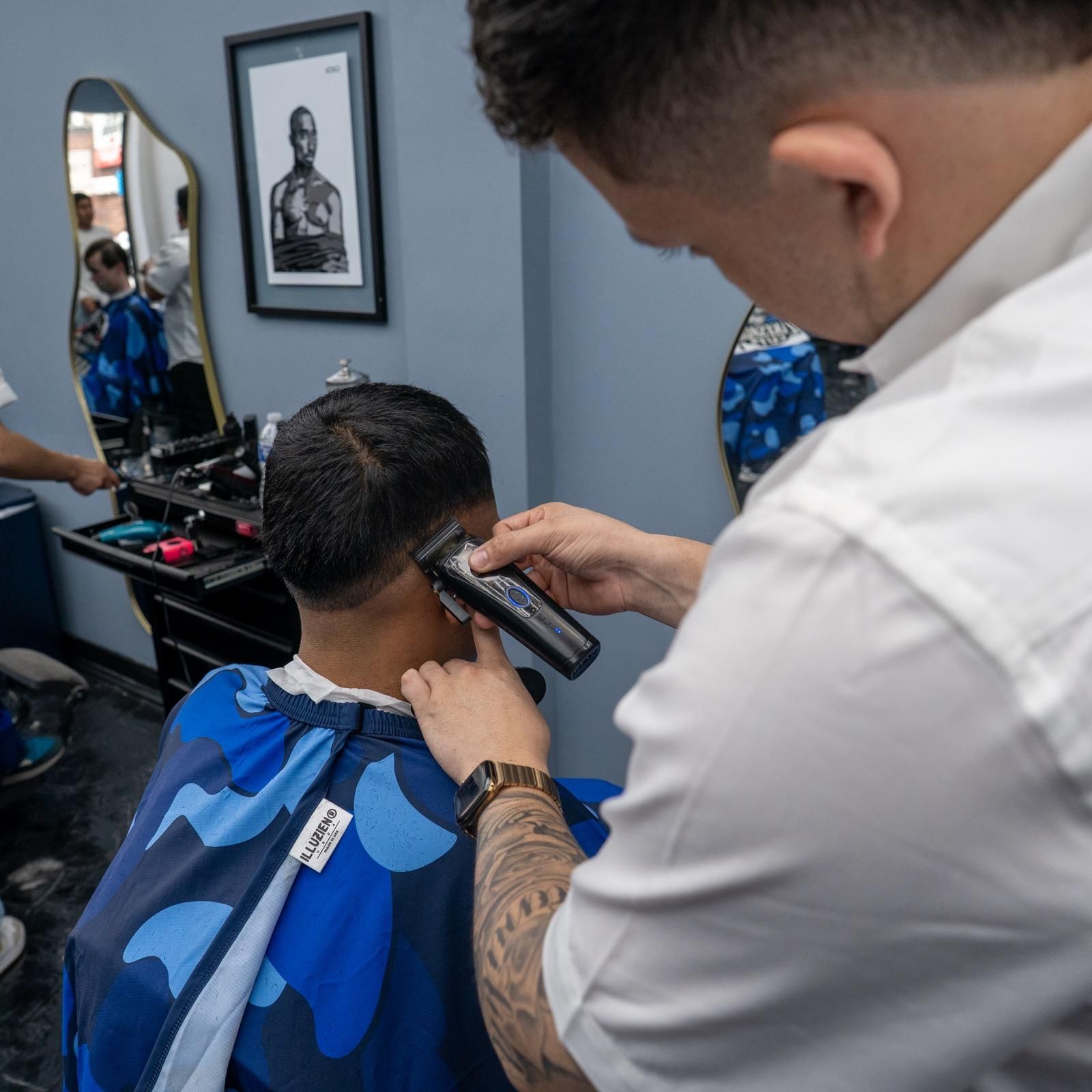 Barber trimming a client's hair with clippers in a blue-toned barbershop.