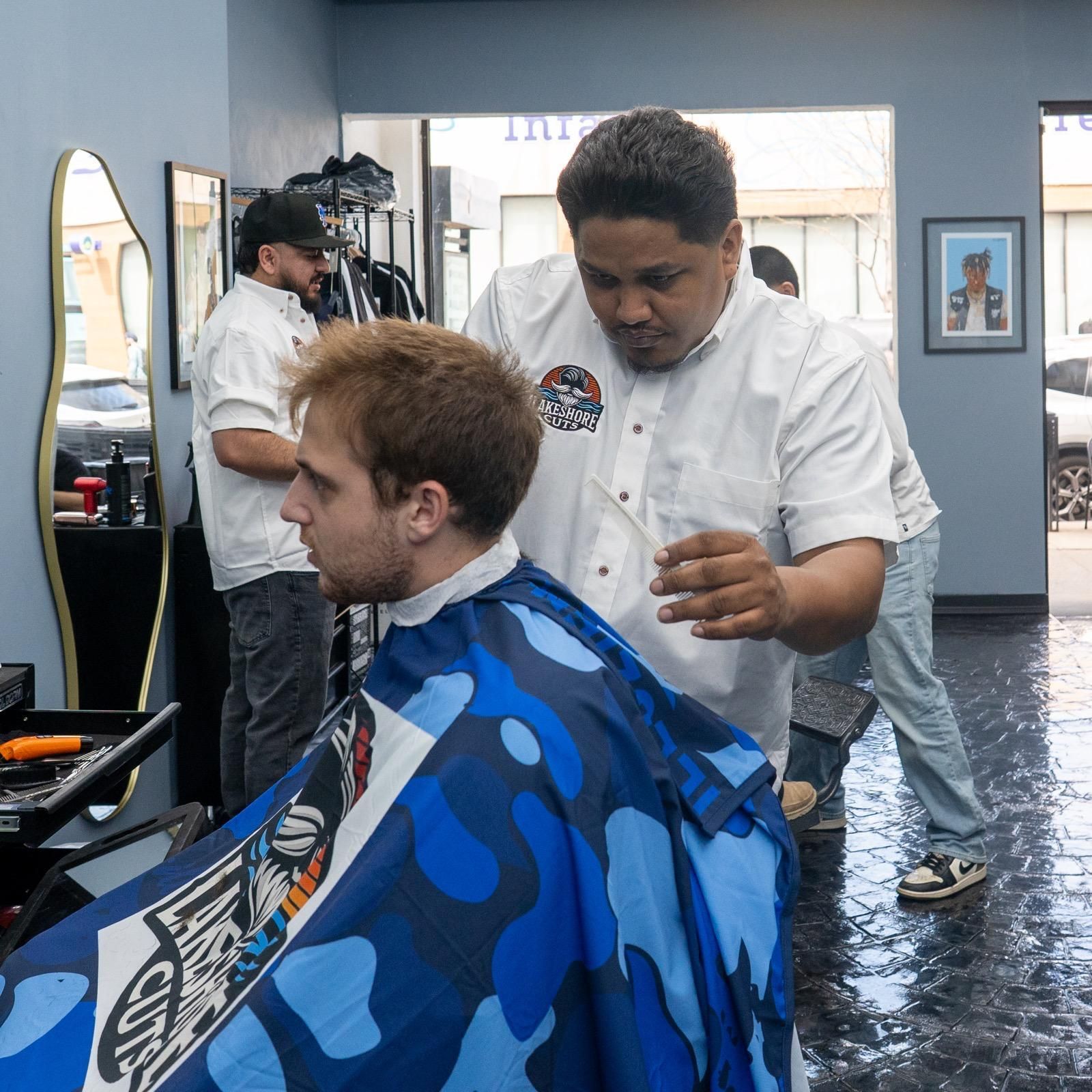 Barbershop haircut in progress, with barber trimming a seated customer in a blue cape.