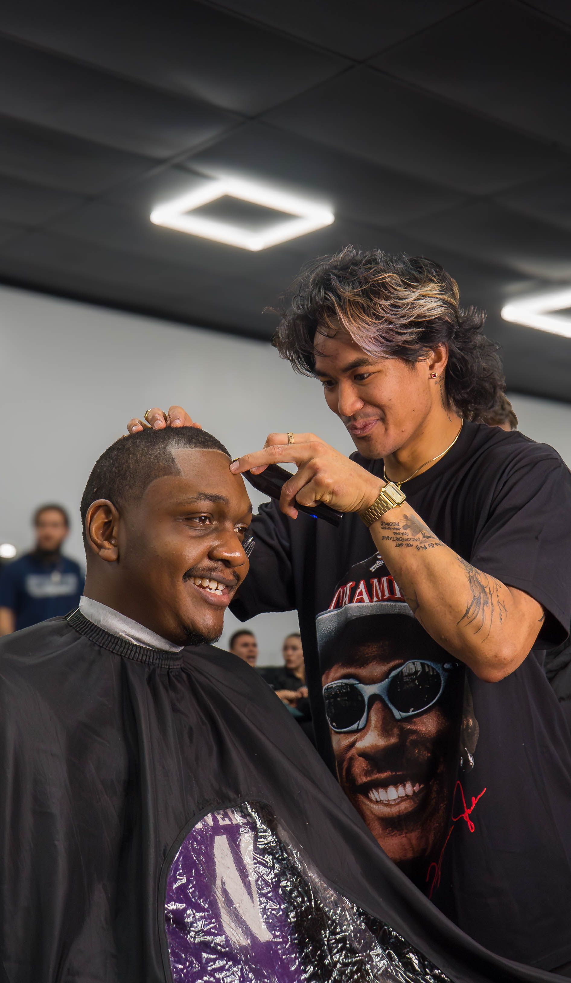Person getting a haircut while another smiles nearby in a barbershop-like setting