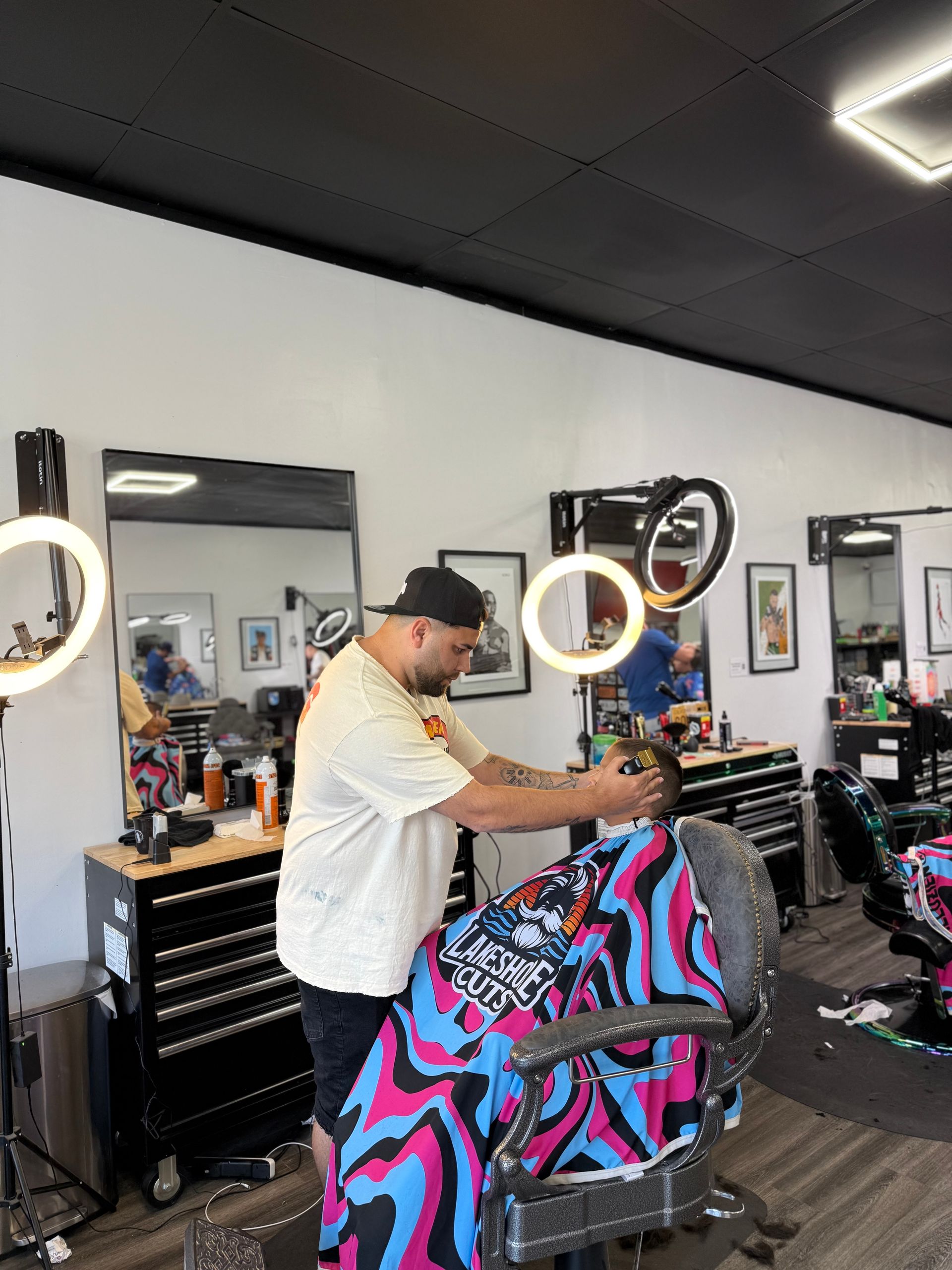 Barber styling a client in a salon chair, with ring lights and colorful cape visible.
