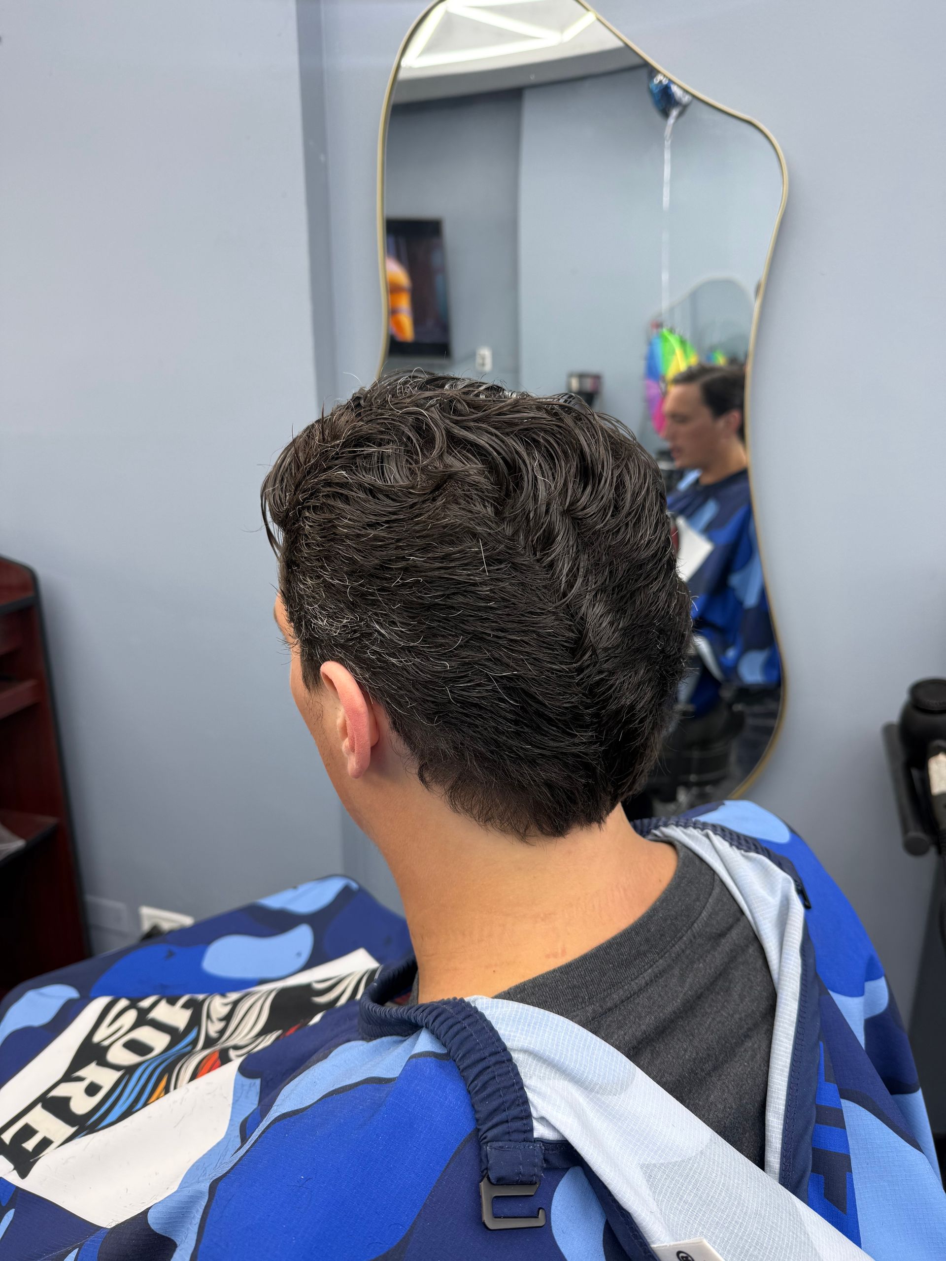 Person with curly hair sitting in a barber chair, seen from behind in a salon mirror.