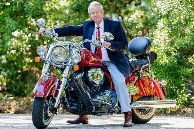 Man sitting on a red vintage motorcycle outdoors, with green foliage in the background