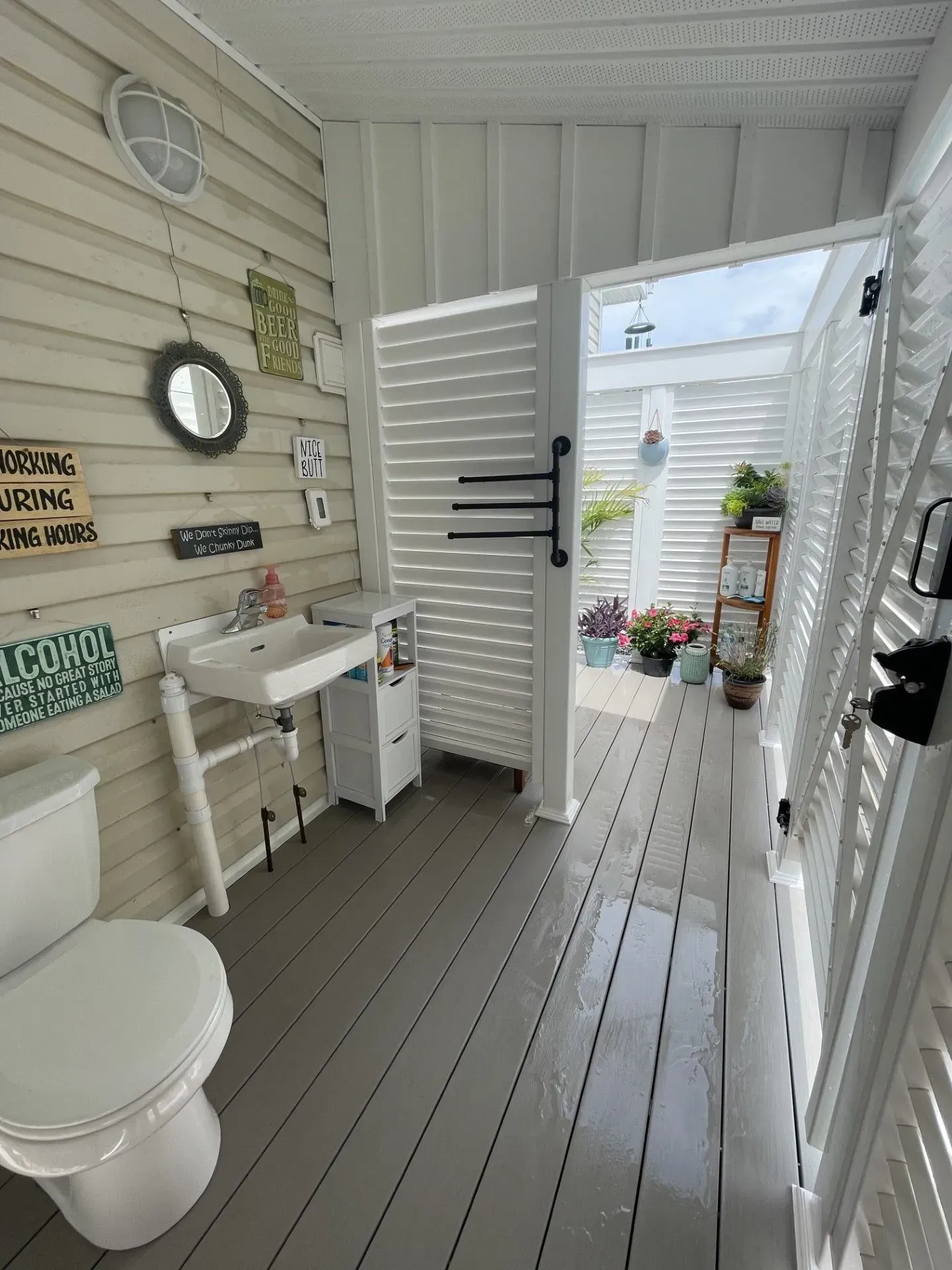 Small white bathroom with sink, toilet, wall mirror, and open shutters leading to a porch.