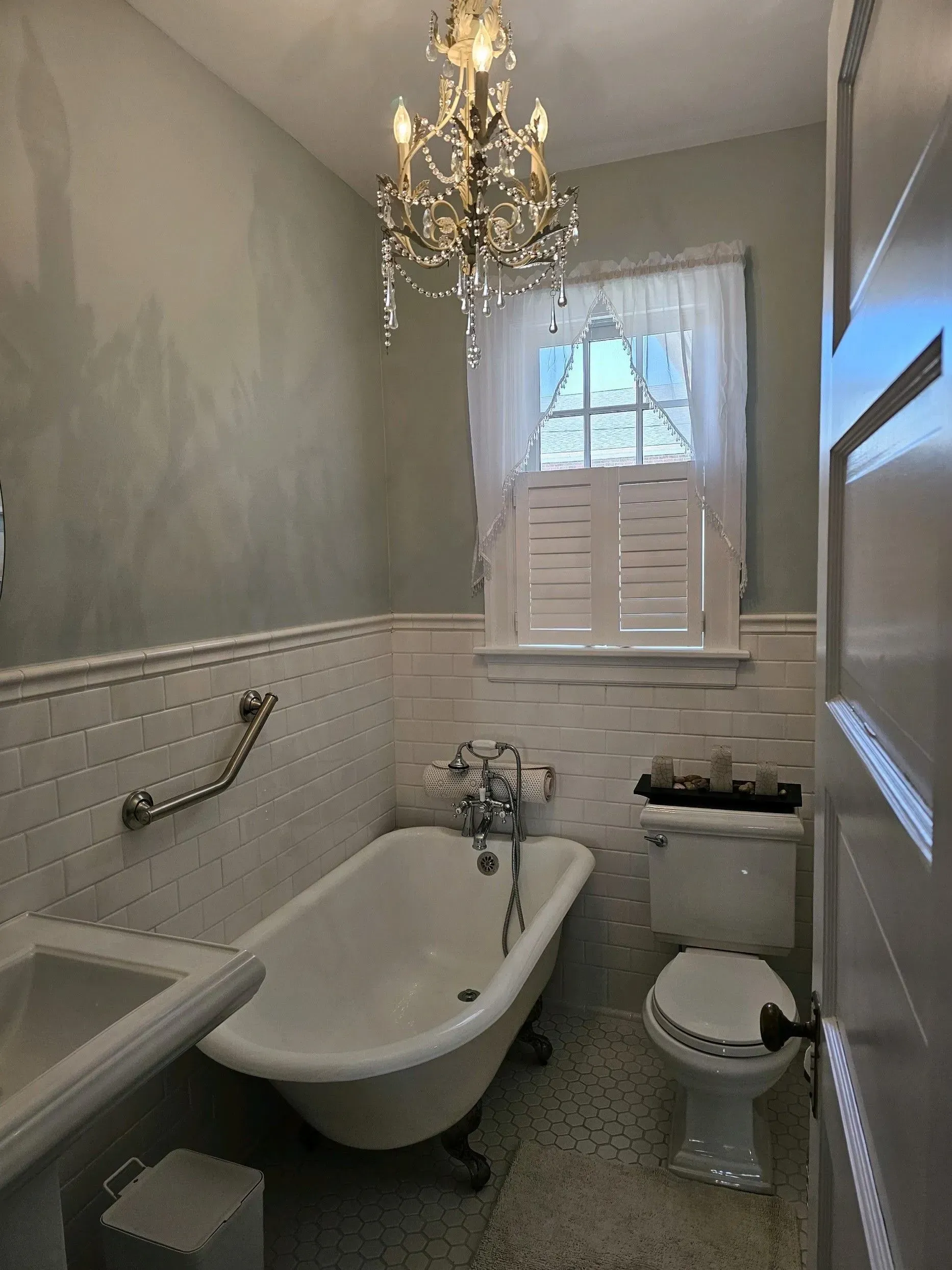 Small vintage bathroom with clawfoot tub, white toilet, sink, chandelier, and a window with shutters.