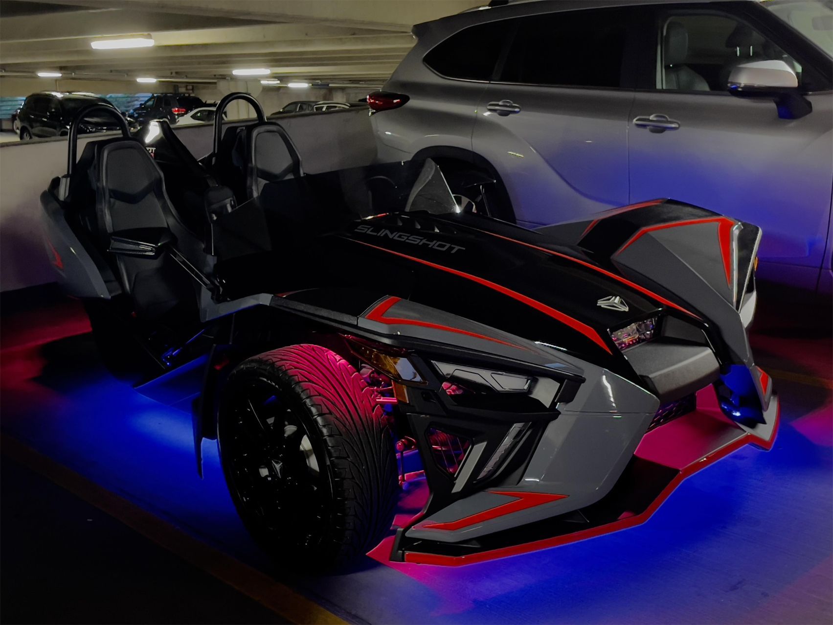 A slingshot is parked in a parking garage next to a car.