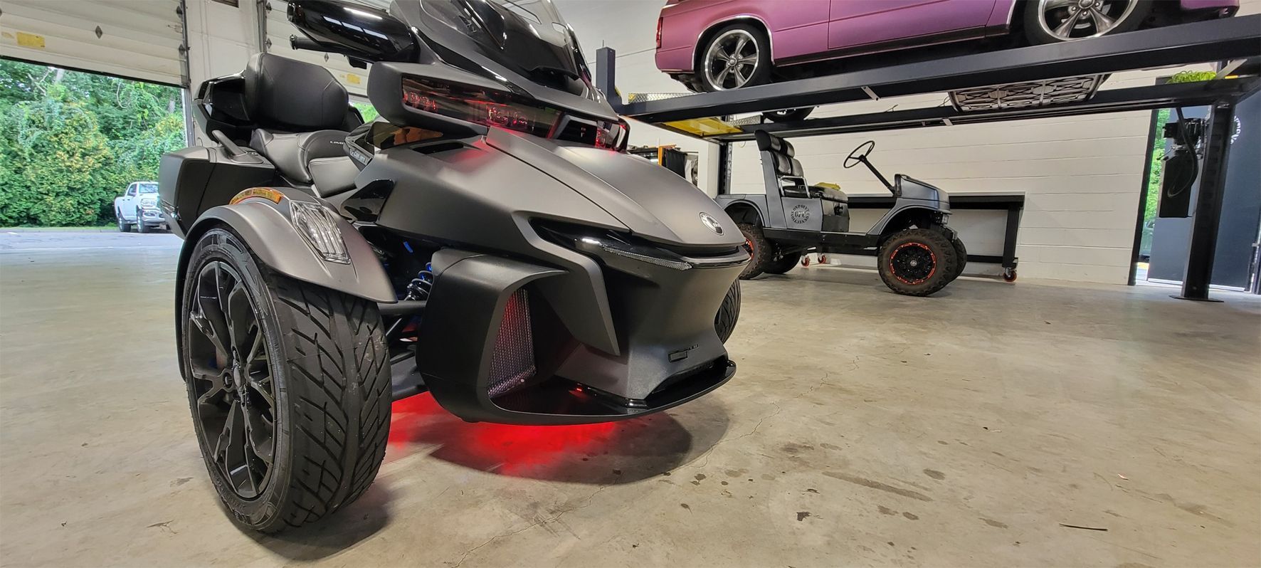 A three wheeled motorcycle is parked in a garage next to a pink car.
