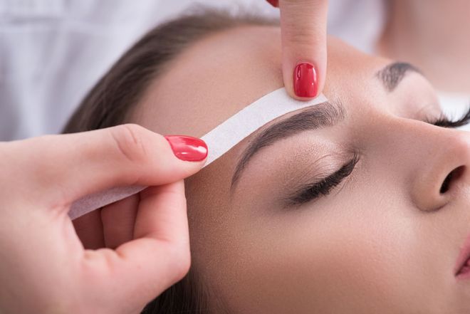 Hand shaping an eyebrow with white wax strip near a closed eye and red-painted nails