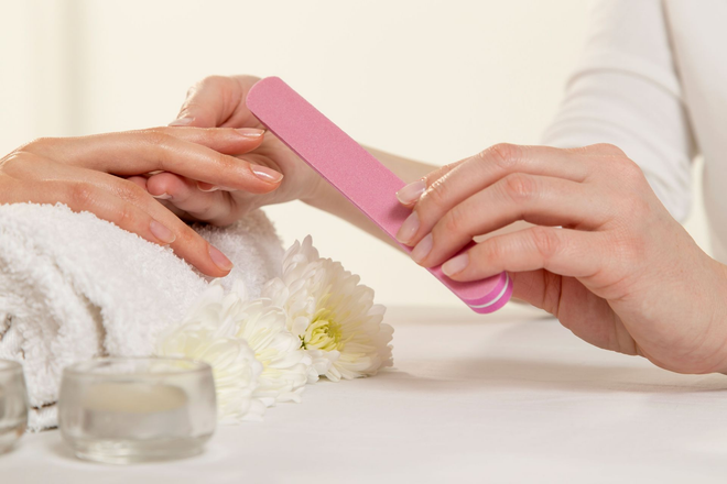 Hands filing nails with a pink emery board beside white flowers on a spa table