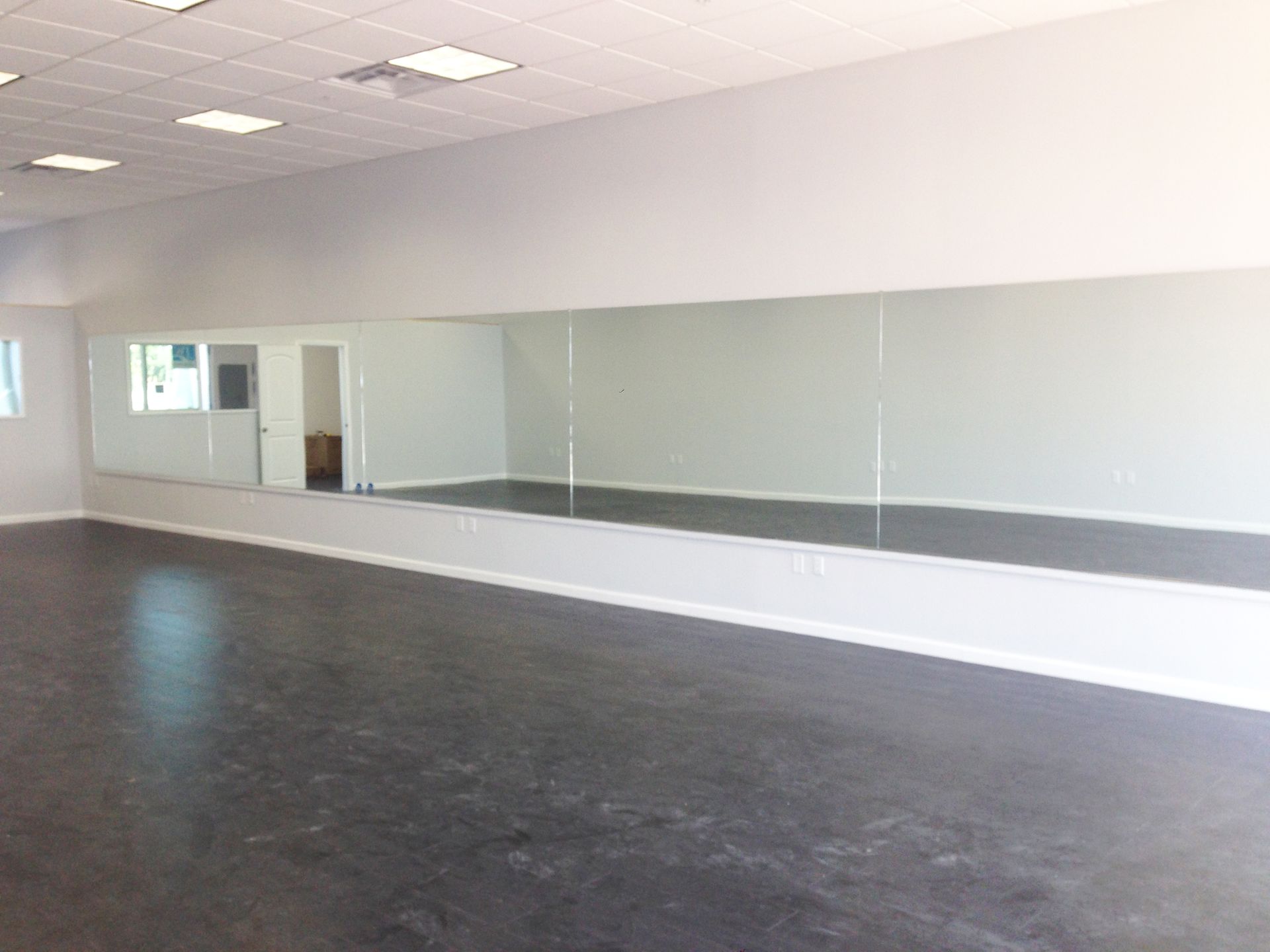 Empty dance studio with a long wall mirror, white walls, and a dark floor.