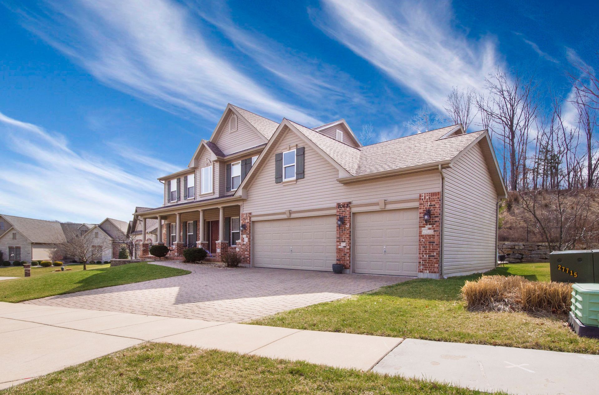 Two-story beige house with a paved driveway, green lawn, and blue sky with wispy clouds.