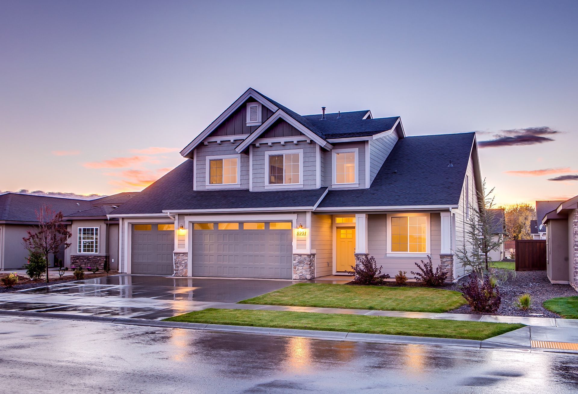 Two-story gray house with a dark roof, two-car garage, and wet driveway at dusk.