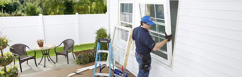 Man in blue cap replacing a window screen on a white house, ladder nearby. Outside, a patio with table and chairs.
