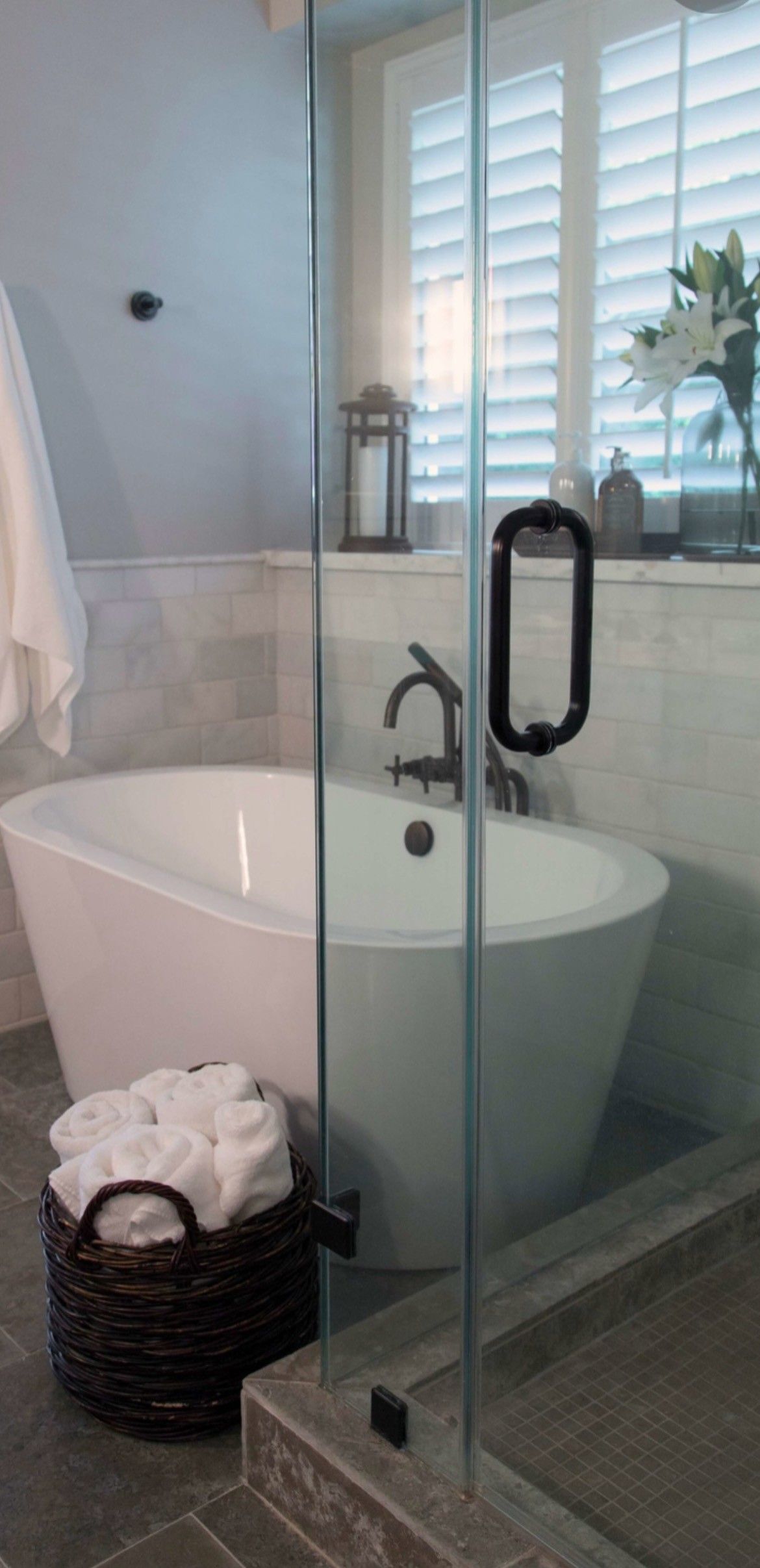 Freestanding bathtub next to glass shower door. White towels in a woven basket. Shutters and flowers in the background.