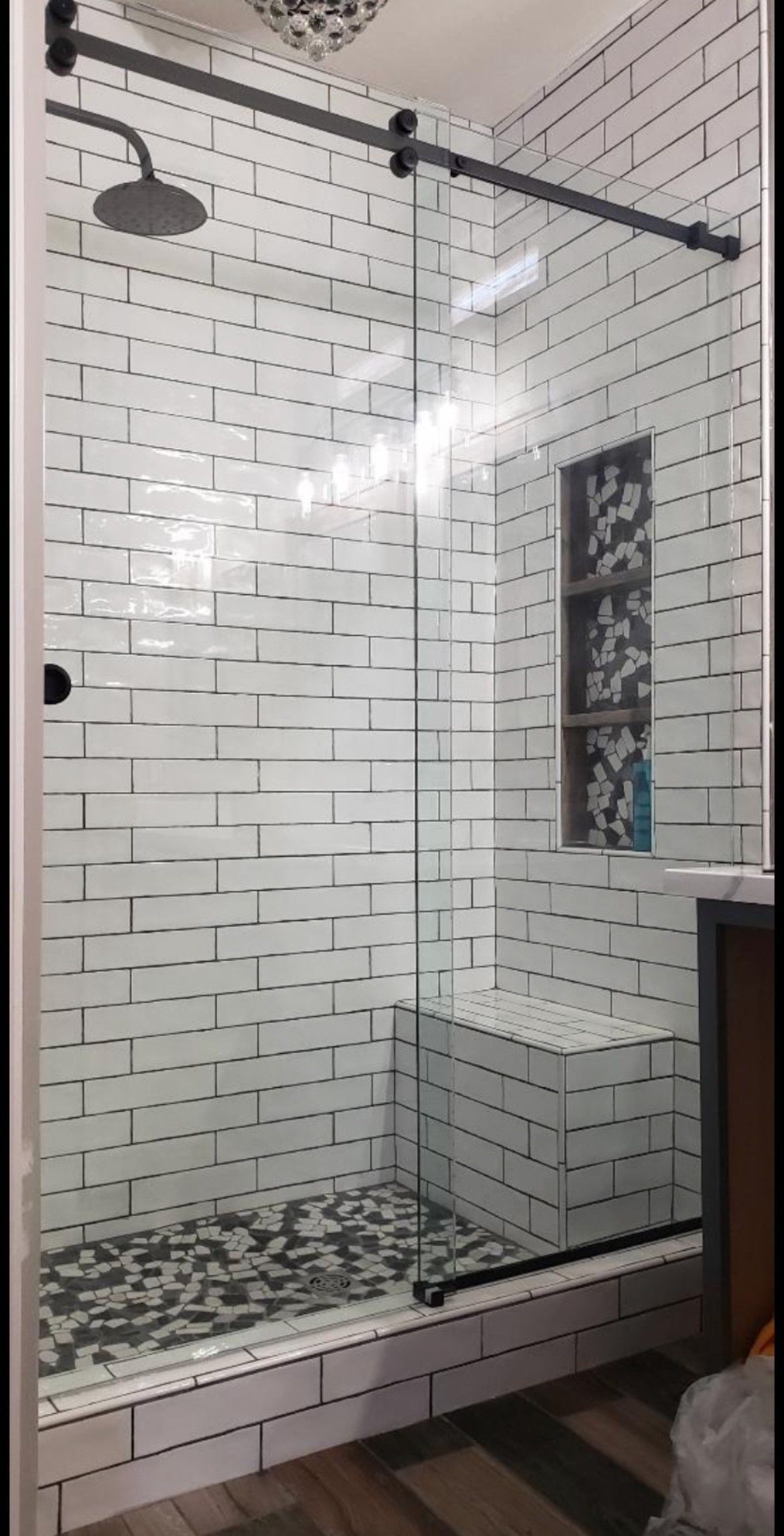 Shower with glass door, white brick-like tiles, black hardware, and a dark pebble floor.