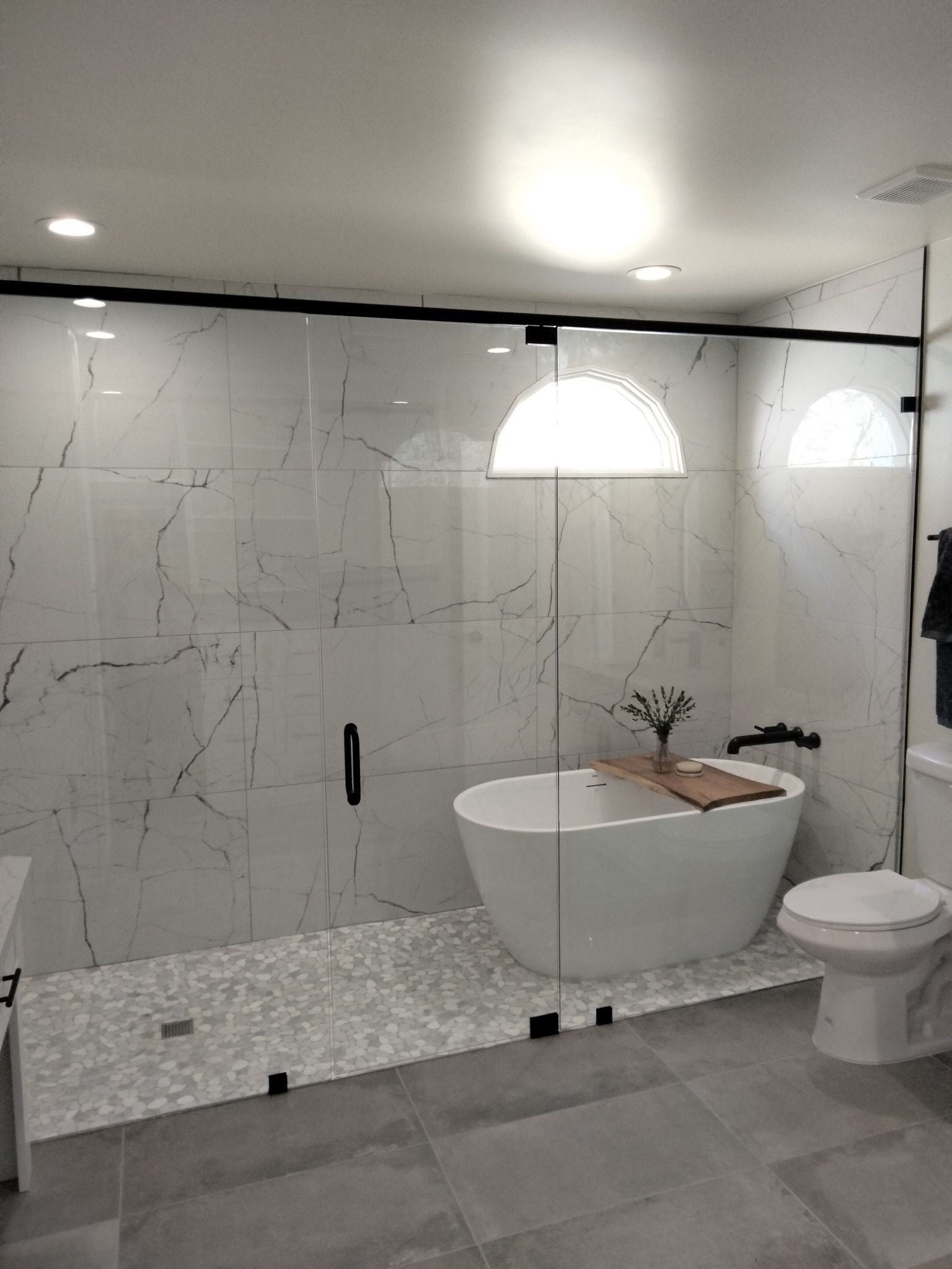 Modern bathroom with marble-look tile, glass shower, freestanding tub, and pebble floor.