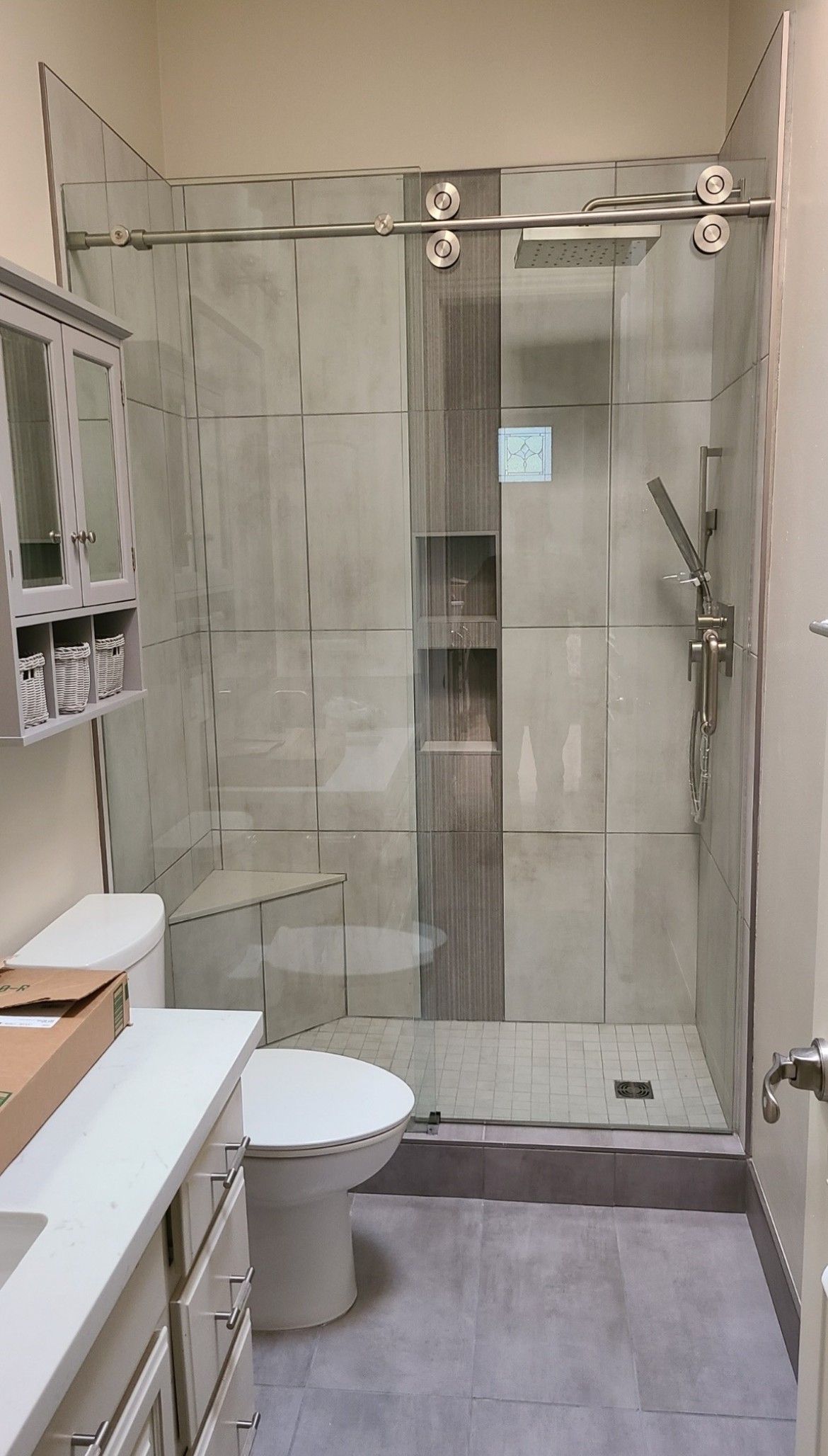 Bathroom with glass shower, gray tile, toilet, vanity, and wall-mounted storage cabinet.