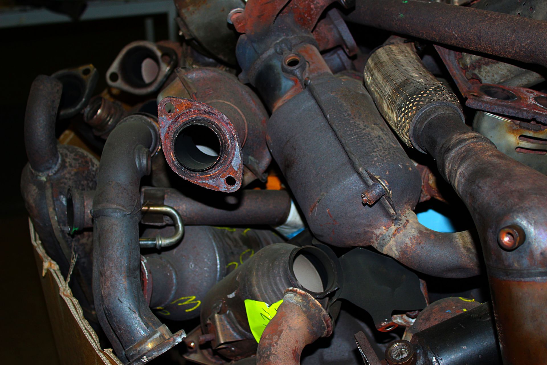 Pile of used catalytic converters in a cardboard box, showing rust and exhaust pipes.