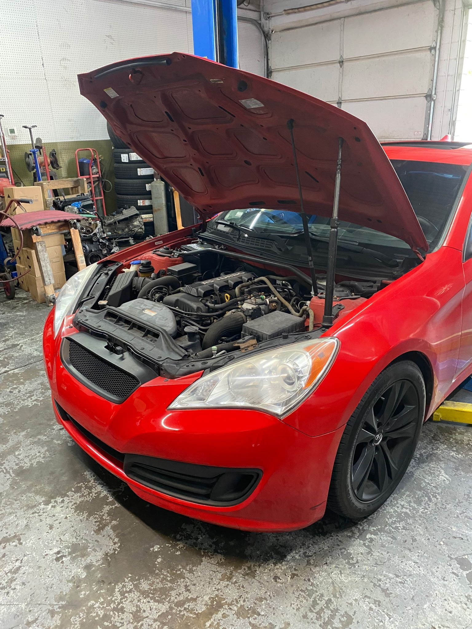 Red car with hood open in a garage, revealing the engine. The vehicle is lifted on a hoist.