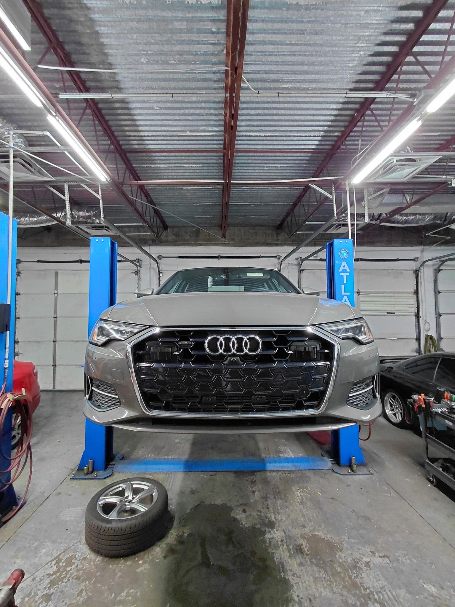 Silver Audi sedan on a blue car lift inside a garage; spare tire on floor.