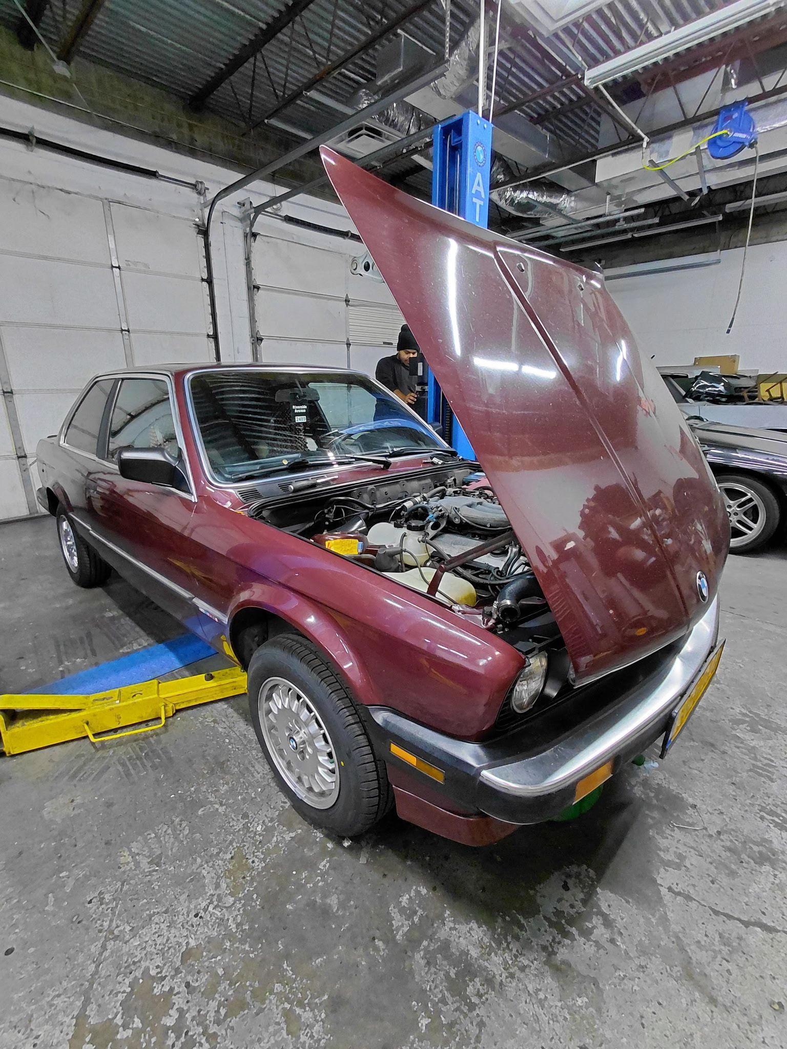 Maroon BMW coupe with hood open in a garage, vehicle on a lift.