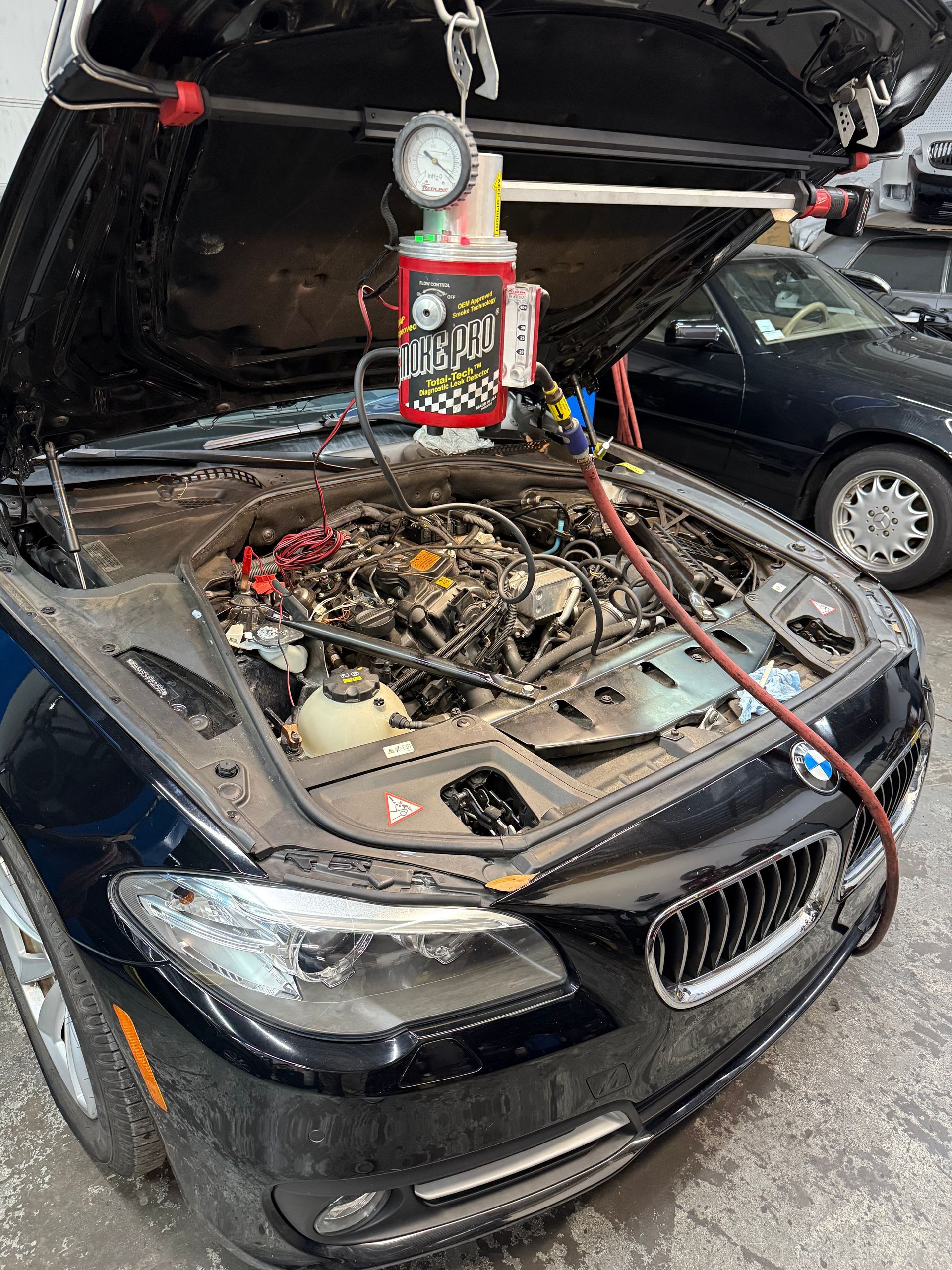 Black car with hood up, engine being worked on, tool attached. Another car in the background.