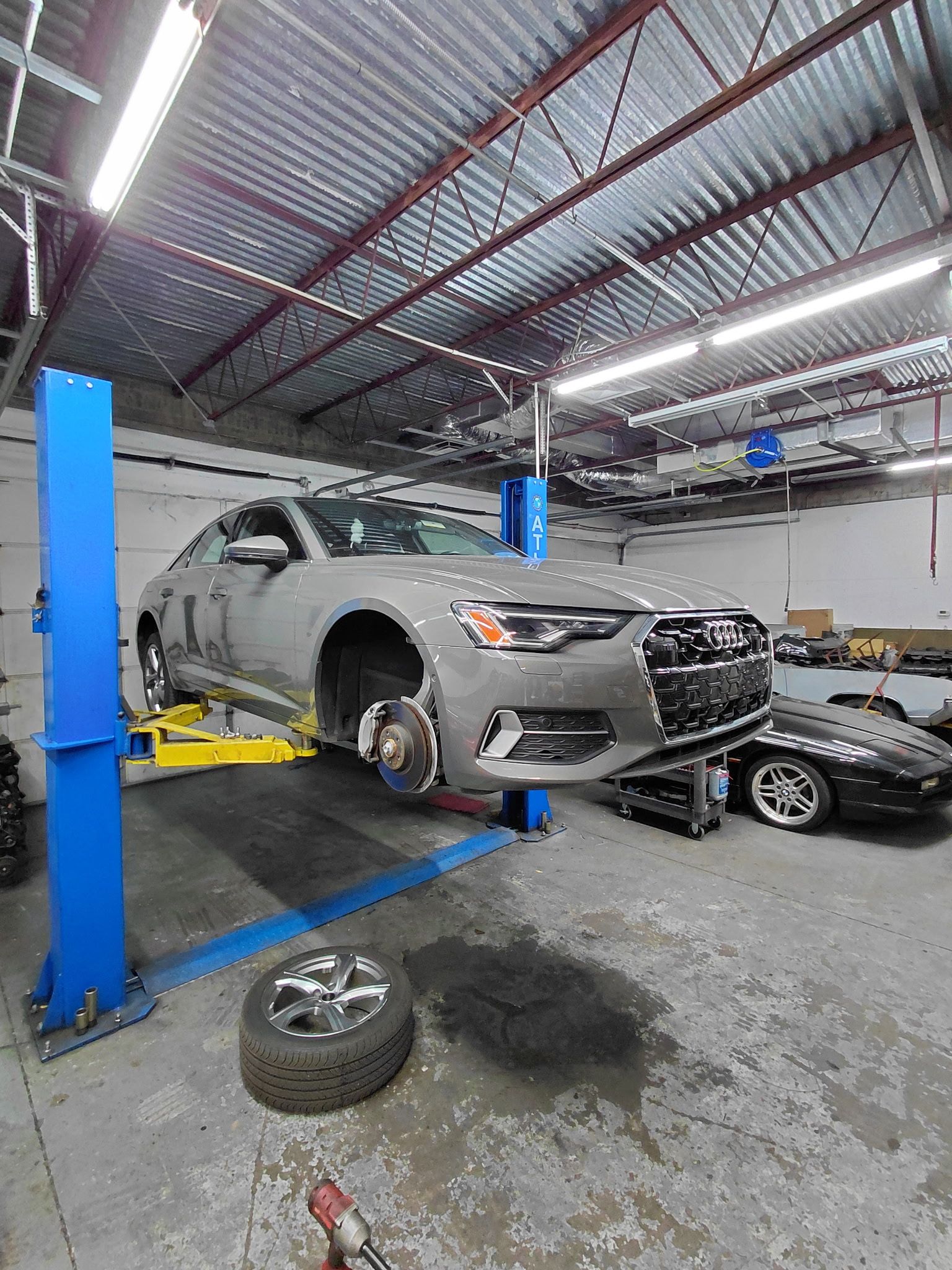 A silver Audi sedan on a lift in a garage, wheel removed, with a tire on the floor.