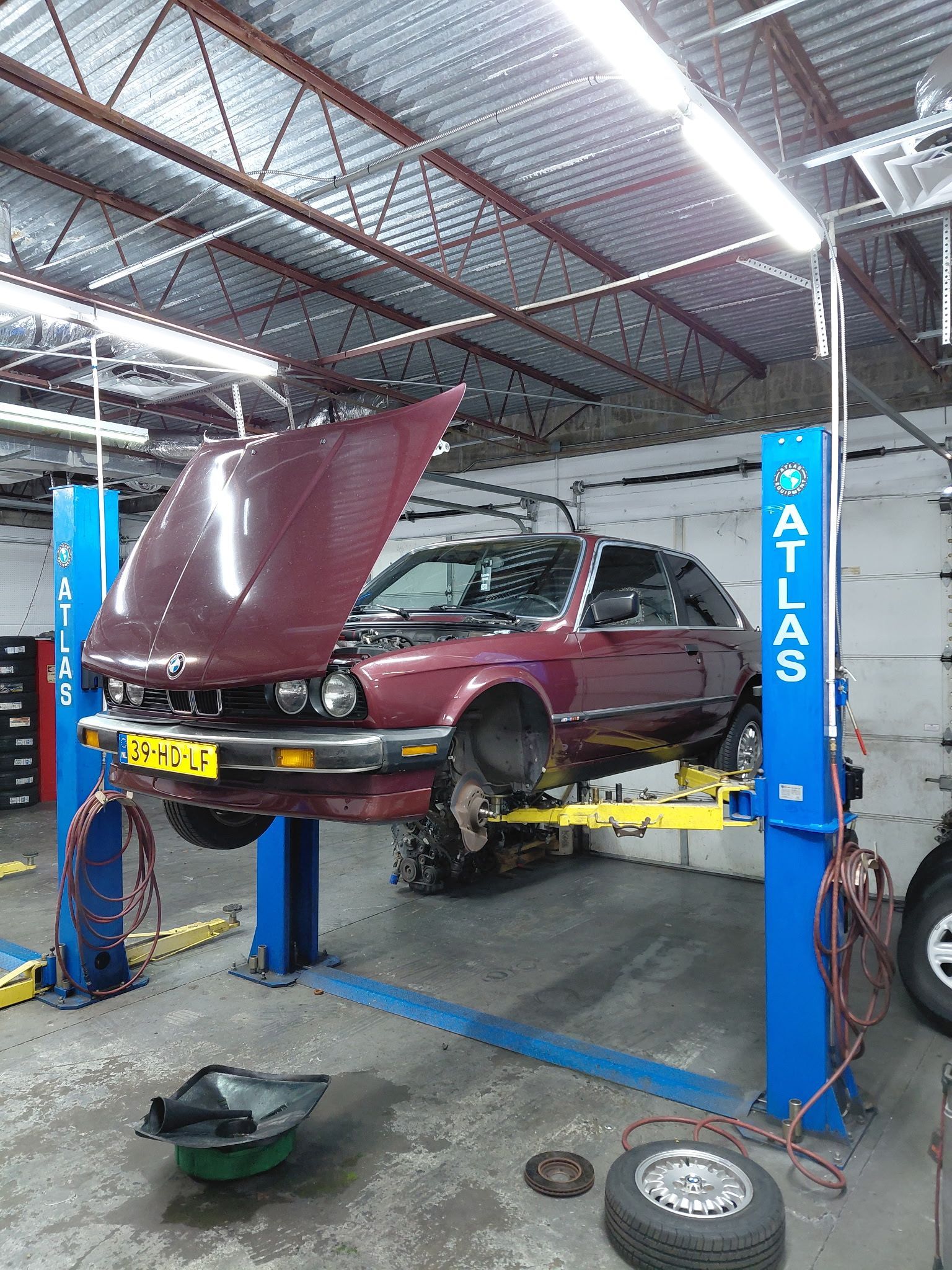 Car raised on a lift in a garage. Maroon vehicle with open hood; wheels removed. Atlas lift posts, gray floor.