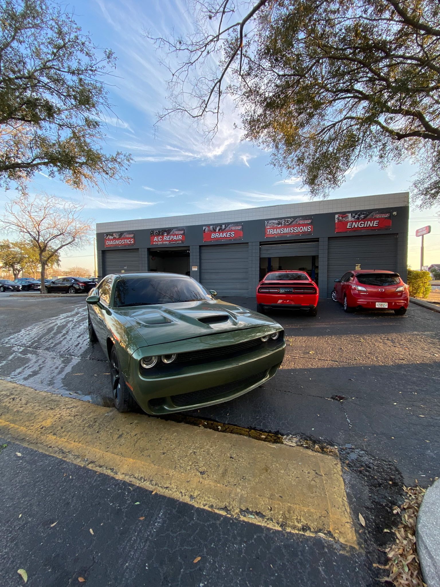 Green Dodge Challenger in front of a car repair shop with two red cars; asphalt, blue sky.
