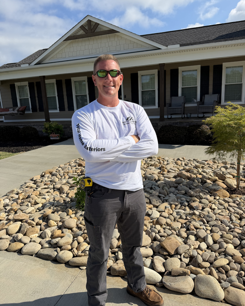 Man standing with arms crossed in front of a house and rock landscaping on a sunny day